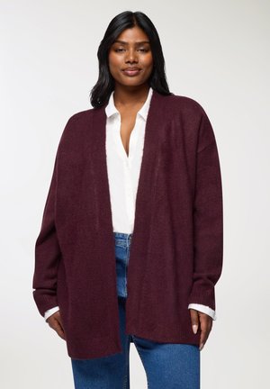 Cardigan in magenta con apertura frontale, maniche lunghe e polsini a coste, indossato sopra una camicia bianca con bottoni e jeans blu. Tessuto morbido.