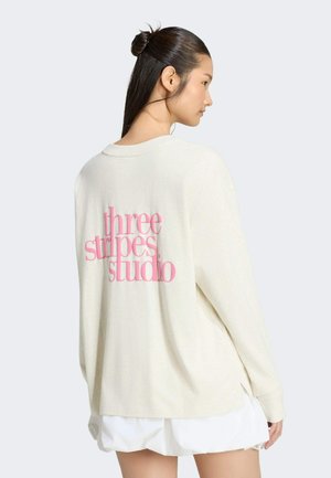 Ung kvinde med mørkt hår samlet op, iført en løs cremefarvet sweater med pink tekst "three stripes studio" på bagsiden over et hvidt skørt.