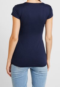 Kvinna klädd i en figurnära mörkblå kortärmad topp och ljusblå jeans, sedd bakifrån mot en neutral bakgrund.