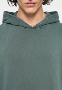 Mørkegrøn ribbet hoodie med en frontlomme og snor i hætten, fremstillet af blødt, struktureret stof. Nærbillede af fronten.