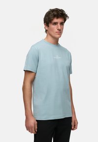 Lyseblå bomulls t-skjorte med rund hals og korte ermer. Har en liten, hvit logo trykket på brystet. Enkelt design.