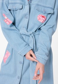 Robe chemise en denim bleu clair avec des designs brodés de rouge à lèvres rose et de beignets, dotée de boutons et d'une ceinture nouée à la taille pour une silhouette bien définie.