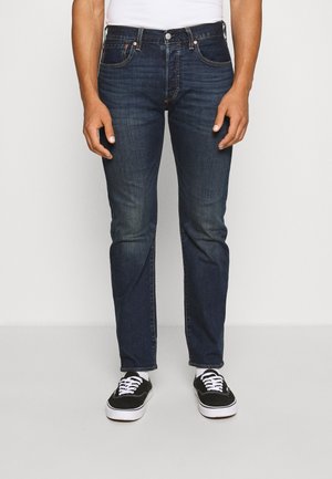 Homme portant un jean slim bleu foncé, des chaussettes blanches et des baskets basses noires avec des lacets et semelles blanches, debout sur un fond clair uni.