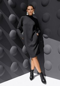 Schwarzes langärmeliges Kleid mit hohem Kragen, seitlichen Taschen und lässigem Schnitt. Kombiniert mit schwarzen Knöchelstiefeln und vor einem strukturierten grauen Hintergrund.