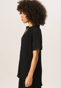Black ribbed shirt with short sleeves, collar, and a buttoned placket. Soft texture and a relaxed fit, featuring a subtle curved hemline.
