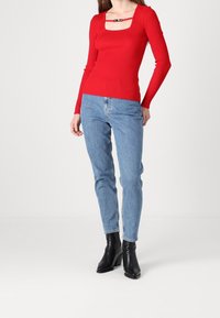 Rotes geripptes Langarmoberteil mit quadratischem Ausschnitt, kombiniert mit hellblauen Jeans. Schwarze stiefeletten mit Absatz runden das Outfit ab.
