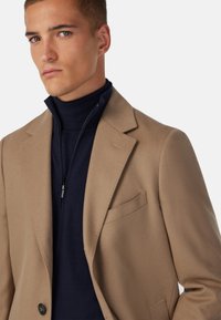 Ljusbrun ullblazer med snedskarv och bröstficka, lager på lager över en marinblå dragkedjeskots. Texturerad tyg med skräddarsydd passform.