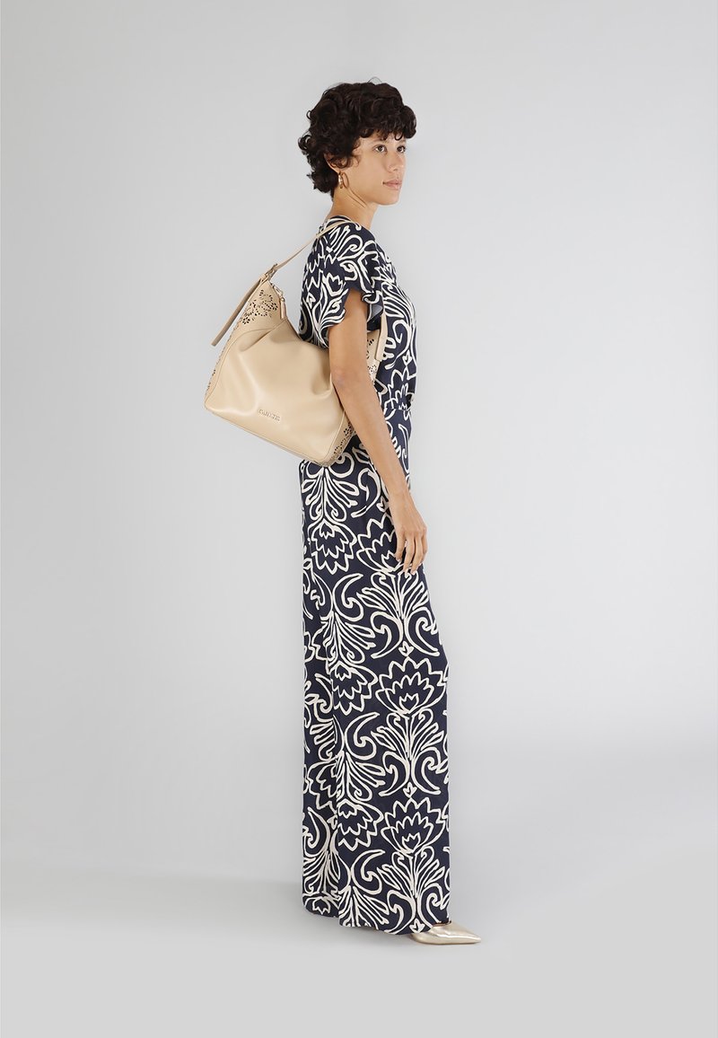 Maxi-jurk met navy basis en wit bloemenpatroon; korte mouwen. Model draagt een beige leren tas met uitsnijdingen en een gladde textuur.