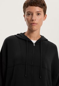 Fekete cipzáras kapucnis pulóver, sima textúrával, húzózsinórokkal és diszkrét varrási részletekkel. Géppel mosható anyag a könnyű ápolás érdekében.