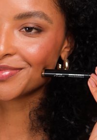 Svart sminkestift merket "Niko" holdt mot en kinn; modellen har krøllete hår og gyldne hoop-øreringer, som viser et naturlig sminkelook.