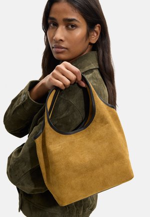 Kobieta w oliwkowej kurtce trzyma musztardową zamszową torebkę z czarnym obszyciem na jednolitym tle.