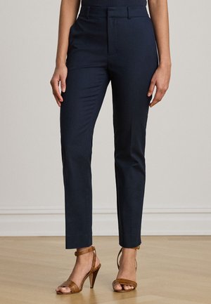 Femme portant un pantalon slim bleu marine foncé et des sandales marron à talons hauts, debout sur un sol en bois contre un mur clair.