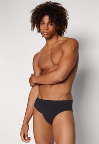 Schwarze Baumwollslips mit glatter Textur, einfachem Design und elastischem Bund mit "SLOGGI"-Logo. Modell posiert mit verschränkten Armen.