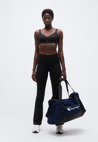 Αθλητική γυναίκα που φοράει μαύρο αθλητικό σουτιέν και κολάν, κρατώντας μια ναυτική και μαύρη τσάντα τύπου duffel της Champion, όρθια μπροστά σε απλό φόντο.