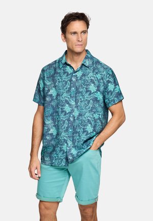 Hombre vistiendo camisa de manga corta con botones y estampado floral en color verde azulado y pantalones cortos de color verde azulado claro enrollados, de pie con una mano en el bolsillo contra un fondo blanco.