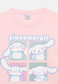 Vaaleanpunainen paita, jossa on neljä sarjakuvamaista valkoista pupuhahmoa, jotka esittävät erilaisia ilmeitä, ja sanan "cinnamoroll" yläpuolella.