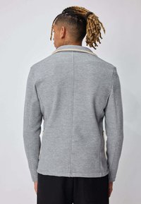 Homme aux dreadlocks blondes attachées en arrière, portant une veste à manches longues gris texturé et un pantalon noir, de dos sur un fond uni.