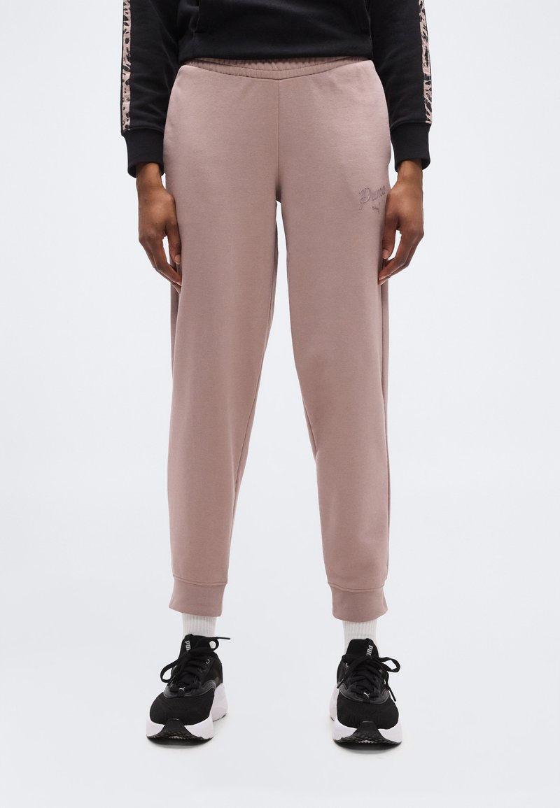 Άτομο που φοράει μοβ παντελόνι φόρμας Puma, λευκές κάλτσες, μαύρα και λευκά αθλητικά παπούτσια Puma, και μαύρο φούτερ με ρίγες λεοπάρ στα μανίκια.