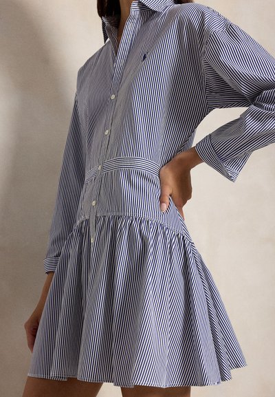 Η γυναίκα φοράει ένα φόρεμα με μπλε και λευκές κάθετες ρίγες, κουμπώνει μπροστά, με μακριά μανίκια και φούστα με πιέτες, το χέρι της ακουμπάει στη μέση.