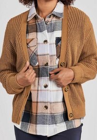 Hnědý pletený kardigan přes kostkovanou košili s velkými knoflíky, náprsní kapsou a odstíny bílé, béžové a šedé.