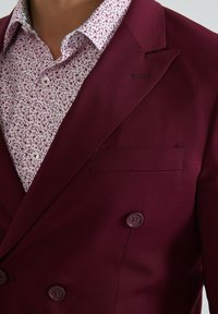 Blazer bordeaux à double boutonnage avec une poche poitrine unique, assorti à une chemise blanche à petit motif floral en rouge et bordeaux.