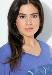 Jonge vrouw met lang donker haar, draagt zilveren oorringen en een blauwe V-hals top met een maan ontwerp.