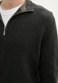 Juodas megztinis su užtrauktuku ant apykaklės, su tekstūruota medžiaga ir kontrastingomis juostelėmis ant kaklo.
