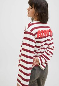 Jonge vrouw met schouderlang donker haar, die wegkijkt. Ze draagt een wit en bordeaux gestreept shirt met lange mouwen, waarop "ATHLETE" op de rug staat.