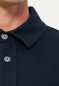 Gros plan d'une personne portant un manteau bleu marine foncé avec deux boutons visibles et un col replié, montrant une partie du cou et du menton.