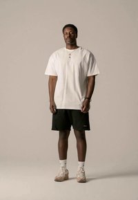 Camiseta de algodão branca, de corte solto, combinada com calções verdes escuros. O modelo veste sapatilhas de cor clara e meias brancas, de pé sobre um fundo claro.