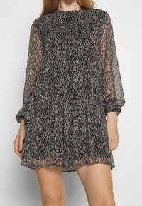 Černé průsvitné šaty s vrstvenou sukní, dlouhými rukávy a jemným leopardím potiskem. Mají vysoký výstřih a knoflíkové detaily.