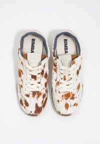 Zapatillas blancas con un patrón moteado marrón, parte superior texturizada similar a piel, cordones crema y forro interior azul. Logo "BIMBA Y LOLA" visible.