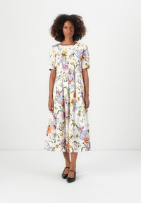 Kukallinen midi-mekko valkoisesta kankaasta, jossa on lyhyet hihat, pyöreä pääntie ja täysimittainen hame, jossa on vaihteleva kukkakuvio. Mustat korot.