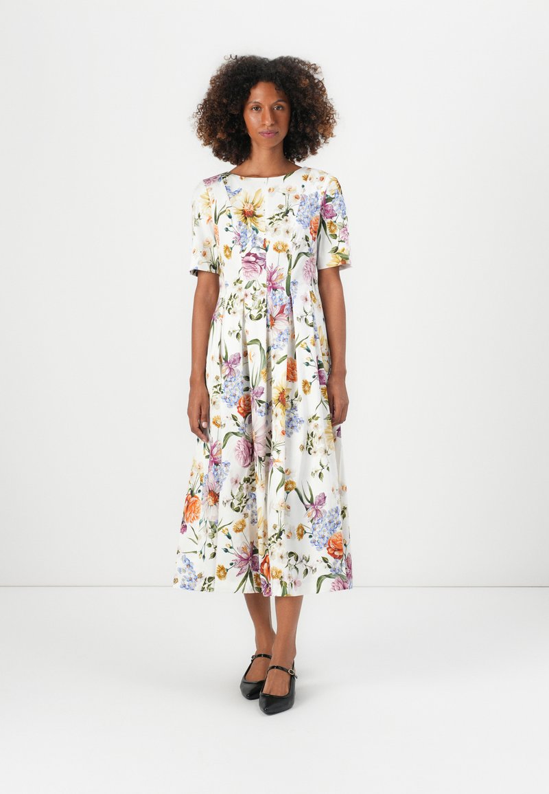 Rochie midi florală din material alb, cu mâneci scurte, guler rotund și fustă amplă cu un model floral variat. Pantofi cu toc negri.