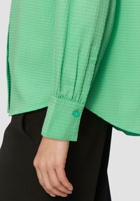 Camicia verde chiaro con texture, maniche lunghe e polsini con bottone, abbinata a pantaloni neri. Il tessuto presenta un motivo sfrangiato.