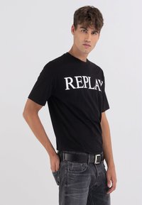 Camiseta de algodón negra con "REPLAY" en letras blancas en negrita. Diseño clásico de cuello redondo, mangas cortas, combinada con jeans grises y un cinturón negro.