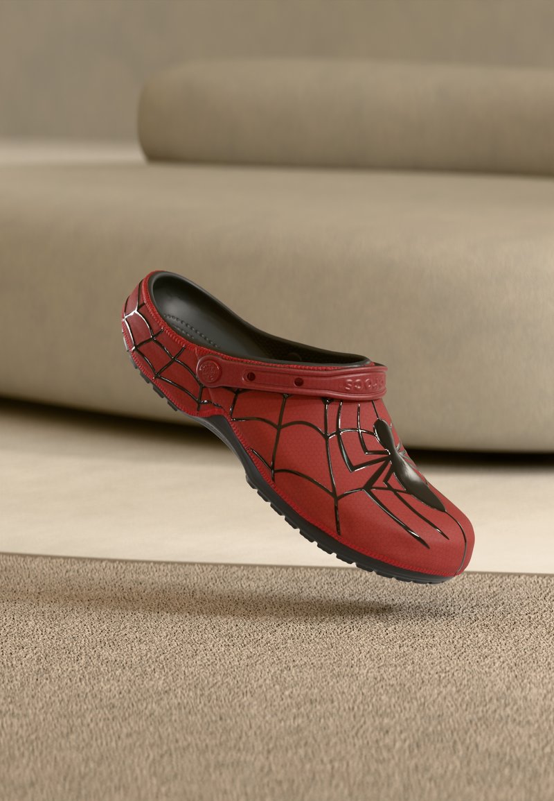 Κόκκινο παπούτσι στυλ κλογκ με μαύρο σχέδιο αράχνης και ιστού, μαύρη σόλα και ρυθμιζόμενο λουράκι φτέρνας, τοποθετημένο σε φόντο μπεζ χαλιού και καναπέ.