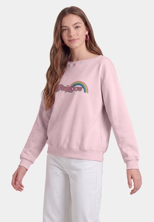 Giovane donna con lunghi capelli mossi che indossa una felpa rosa con una grafica arcobaleno e pantaloni bianchi, in piedi con le braccia leggermente aperte.