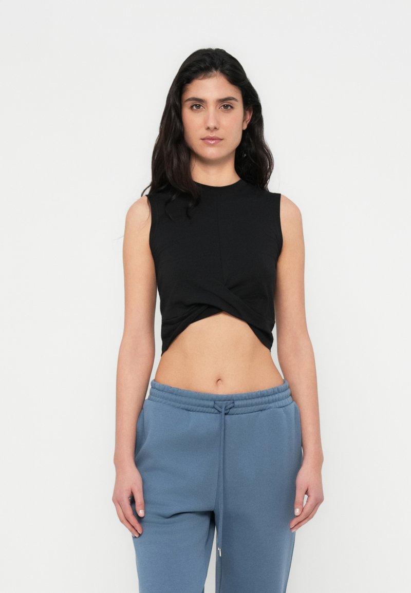 Μαύρο αμάνικο τοπ με σχεδιασμό με κόμπο μπροστά. Συνδυάζεται με μπλε φόρμες, παρουσιάζοντας ένα casual στυλ.