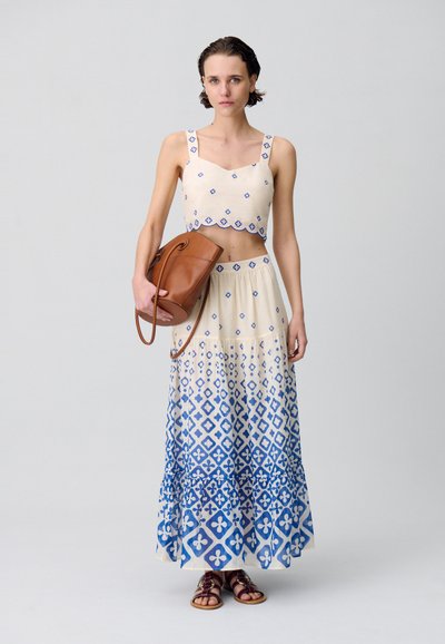 Top corto de crema claro con patrón de diamantes azules y bordes scalloped, combinado con una falda fluida de capas en azul y blanco. Bolso marrón y sandalias.