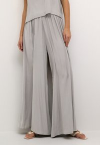 Pantalons larges gris clair en tissu lisse et fluide, avec des plis à la taille et une coupe décontractée. Des sandales sont portées.