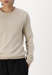 Beige gebreide trui met een textuurpatroon, ronde halslijn en ribgebreide manchetten en zoom. Gekozen boven zwarte joggingsbroek met koord.
