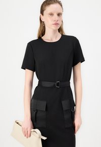 Black dress with short sleeves, round neckline, belted waist, and two front pockets. Model holding a beige clutch. Smooth texture.