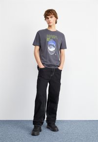 T-shirt graphique NASA gris avec imprimés verts et bleus, associé à un pantalon noir avec des coutures contrastées et des chaussures noires.