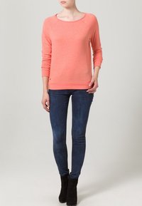 Korálová bavlněná mikina s dlouhými rukávy a kulatým výstřihem, kombinovaná s tmavě modr�ými úzkými džínami a černými kotníkovými botami.