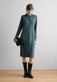 Robe verte à manches longues et fente latérale, en tissu lisse. Associée à des bottes noires à talons mi-mollet et une pochette noire.