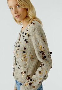 Beige gebreide cardigan met ronde pailletten in goud en zwart, met een ronde hals, lange mouwen en voortassen. Geweven stof.