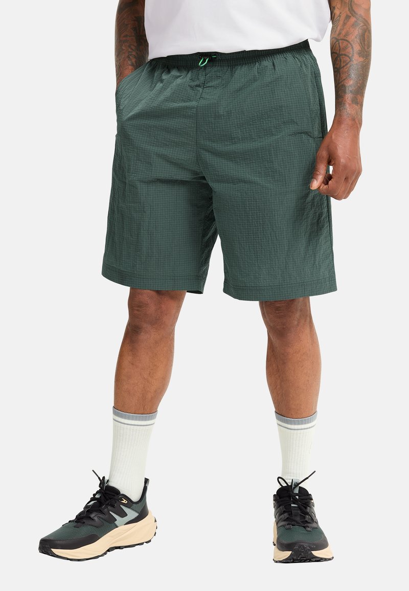 Green shorts made of lightweight fabric with a textured pattern, elastic waist, and side pockets, paired with black and beige sneakers.