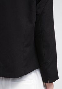 Manche de blazer noir sur mesure sur un pantalon blanc texturé avec poche arrière, vue de profil sur un fond clair.