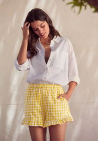 Weißes Leinenhemd mit hochgekrempelten Ärmeln, kombiniert mit gelben Karo-Shorts mit Rüschensaum und blumigen Akzenten.